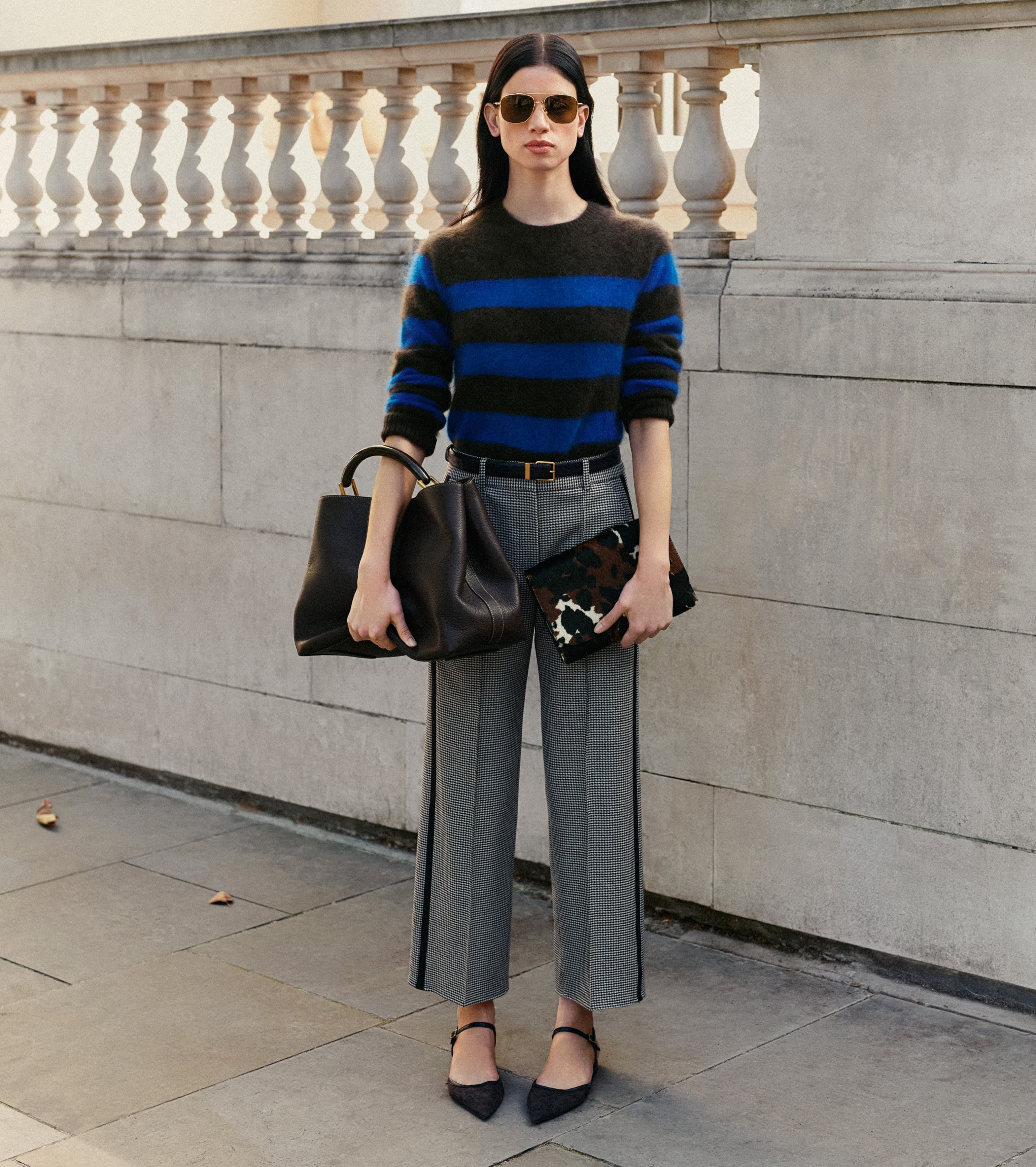 A woman stood in front of a stone wall wearing a striped jumper and carrying two handbags.