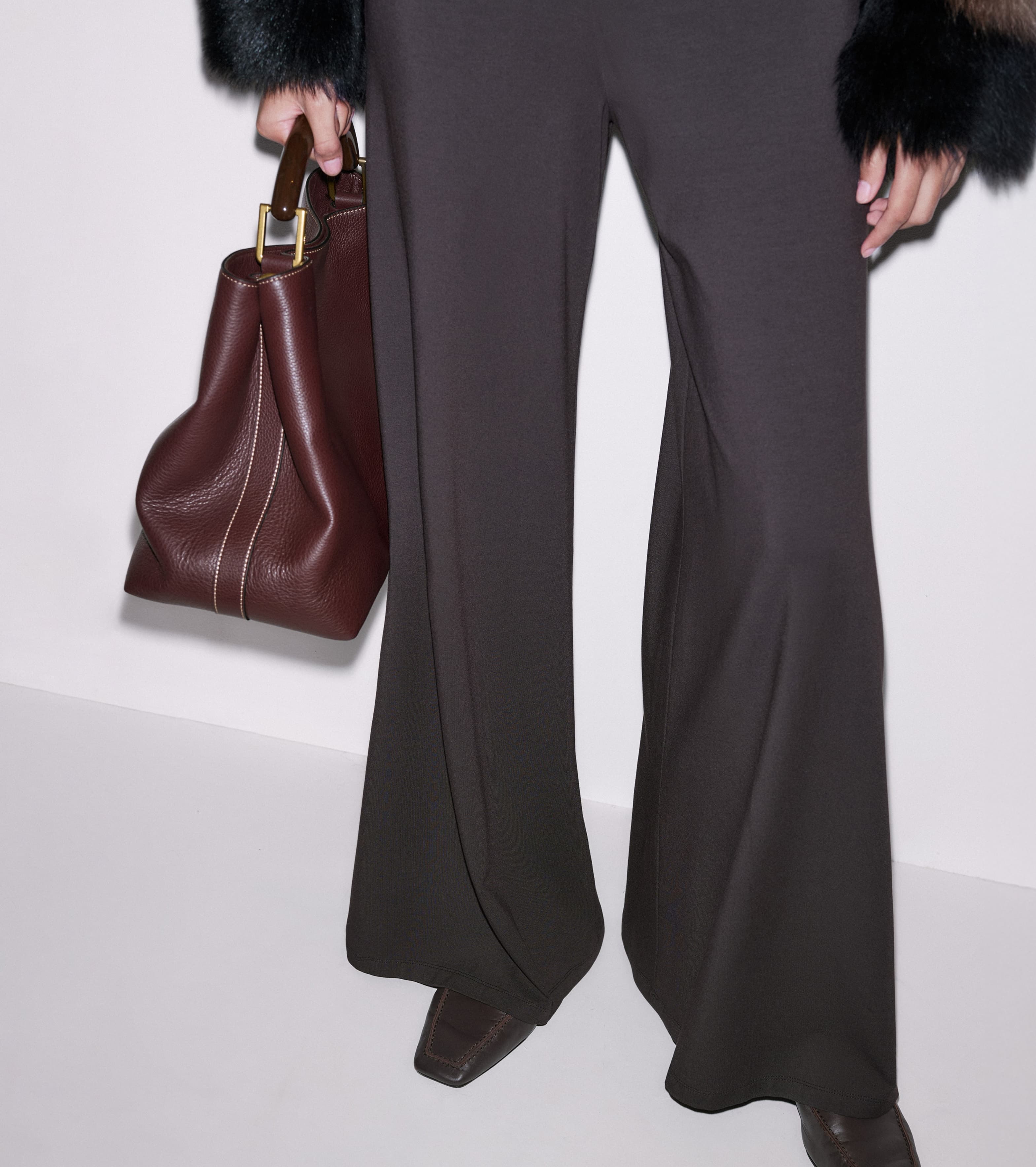 Close up shot of a woman's legs wearing brown trousers and a burgundy leather handbag.