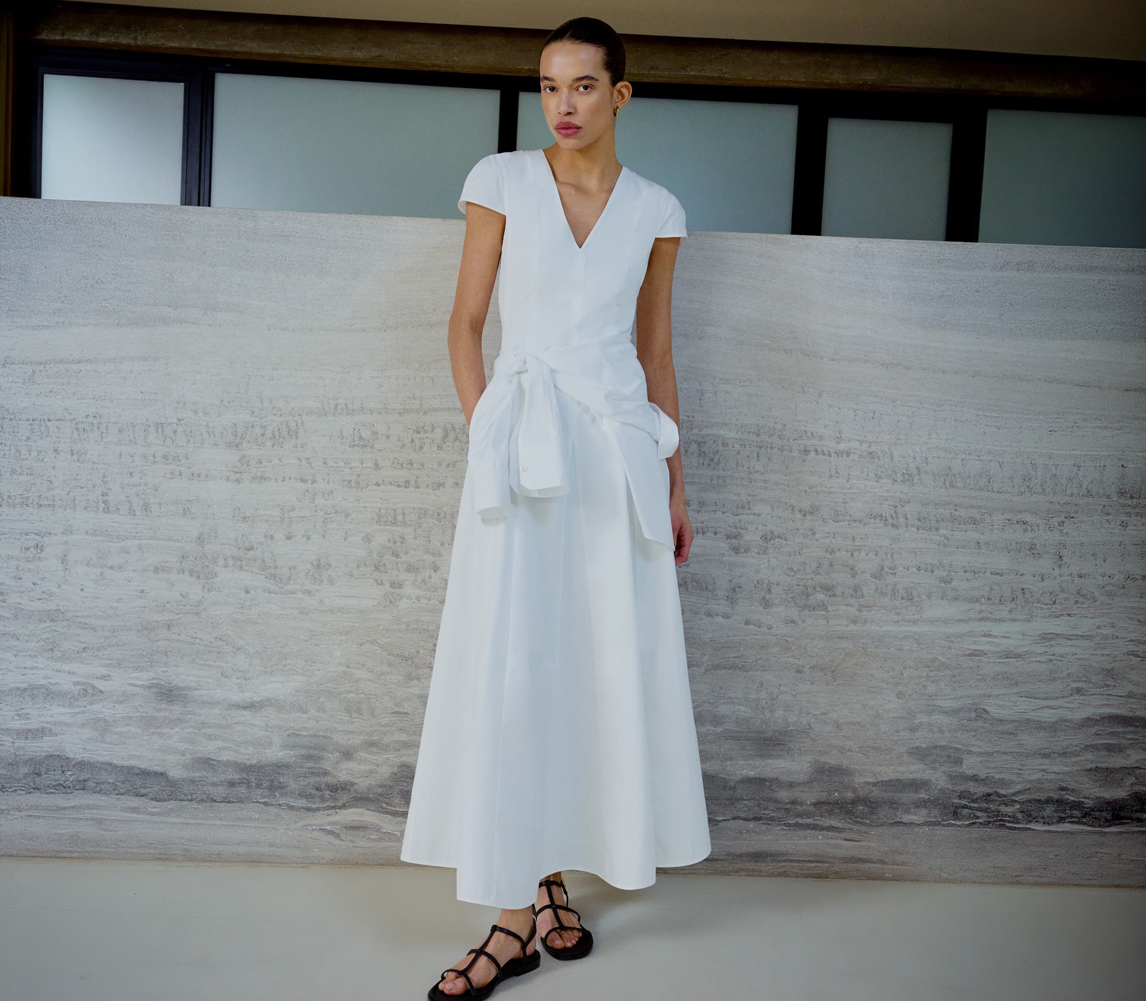 Woman stood in front of a concrete wall in a long white dress with a white jumper tied around her waist.