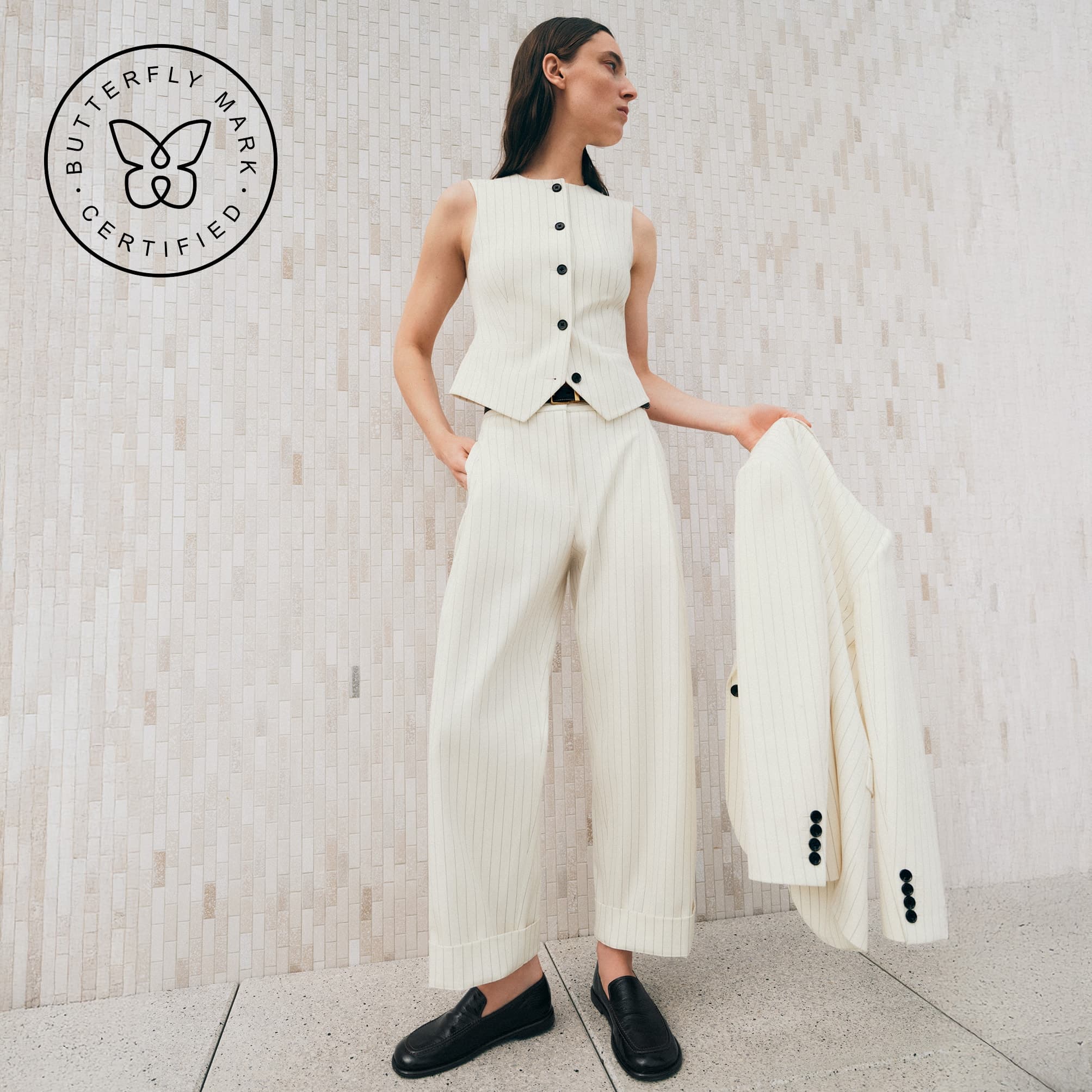 Model standing holding white blazer wearing matching white trousers and a waistcoat with black loafers