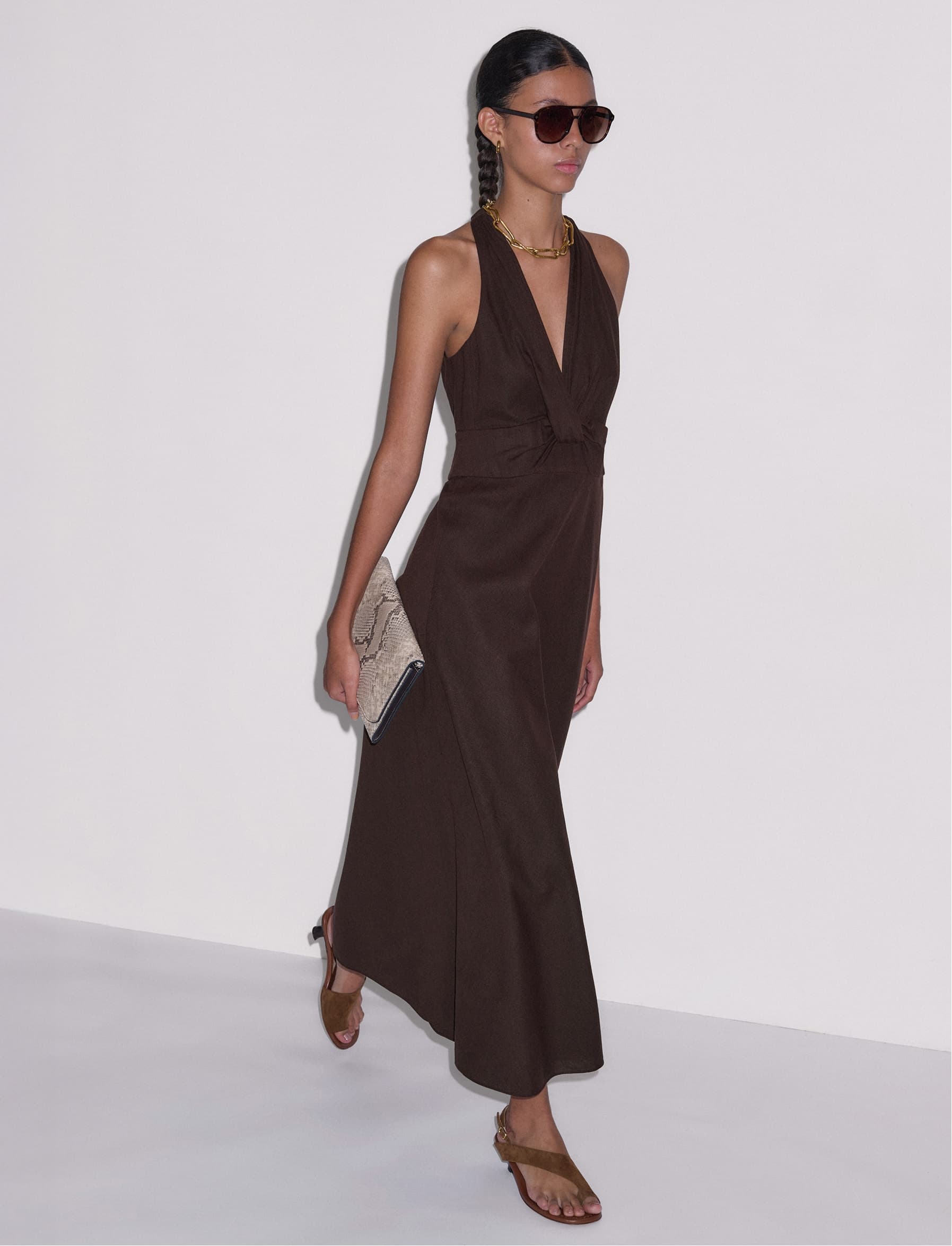 Model walking shot, wearing sunglasses, a brown dress and holding a snake print clutch bag.