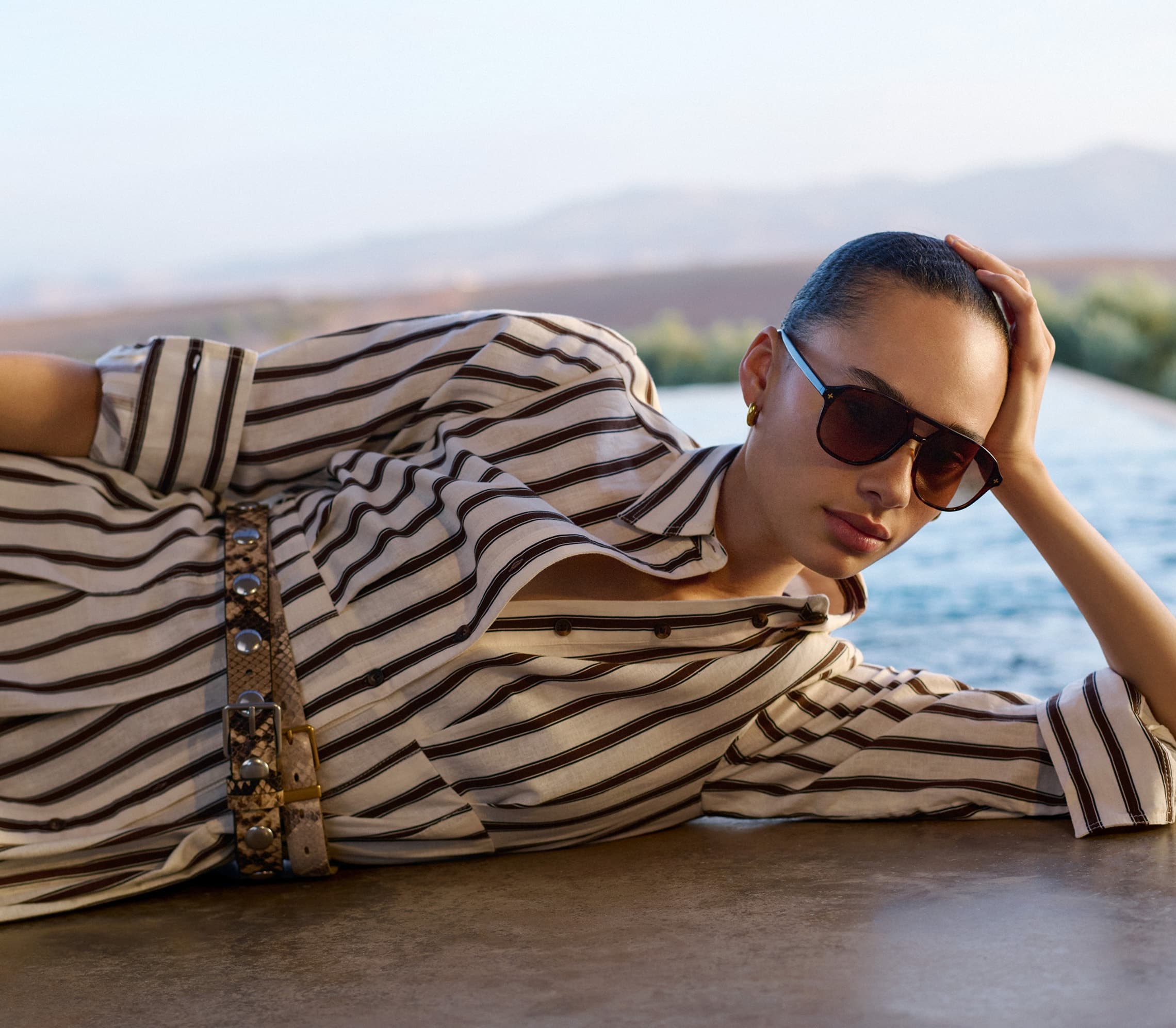 Woman wearing sunglasses and a striped dress lying near a pool with a landscape background.