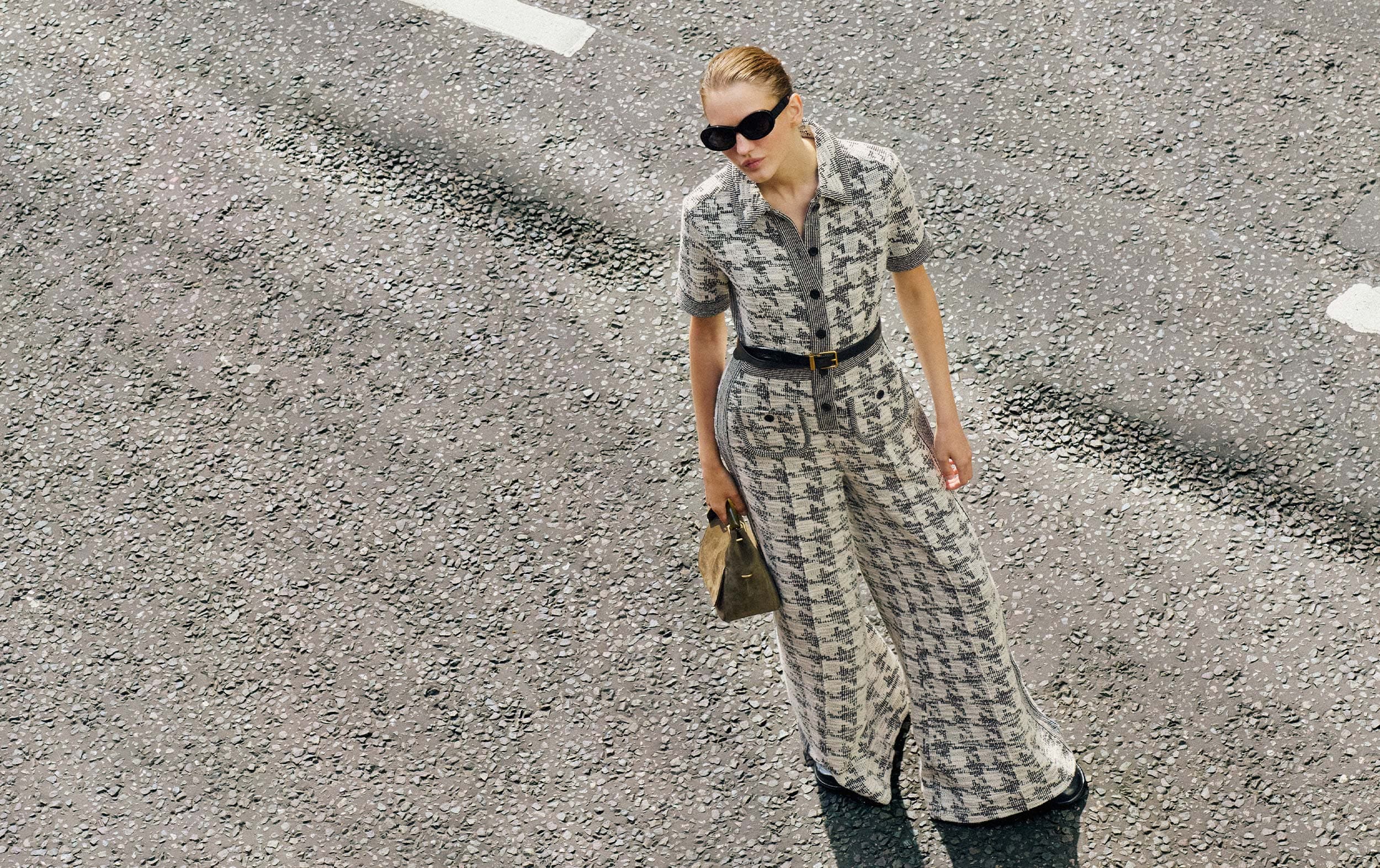 Model wearing black and white houndstooth jumpsuit standing on concreate looking up with sunglasses and holding a handbag