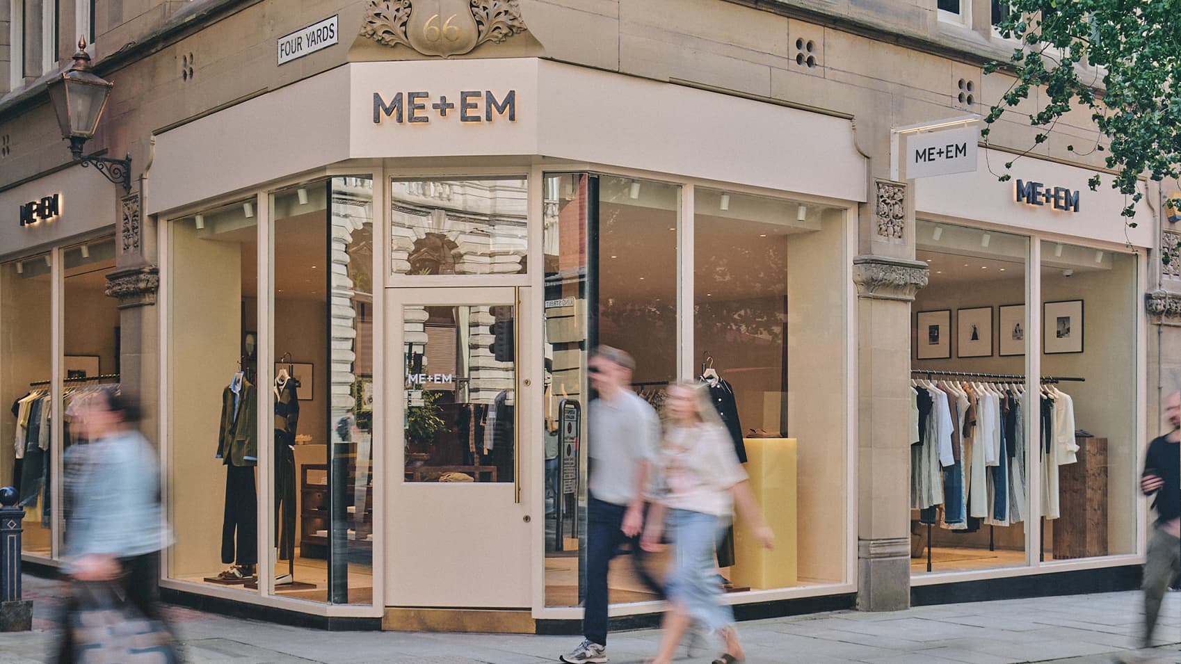 ME+EM Manchester store front with white bricks and clothes hanging in windows with people blurred walking past