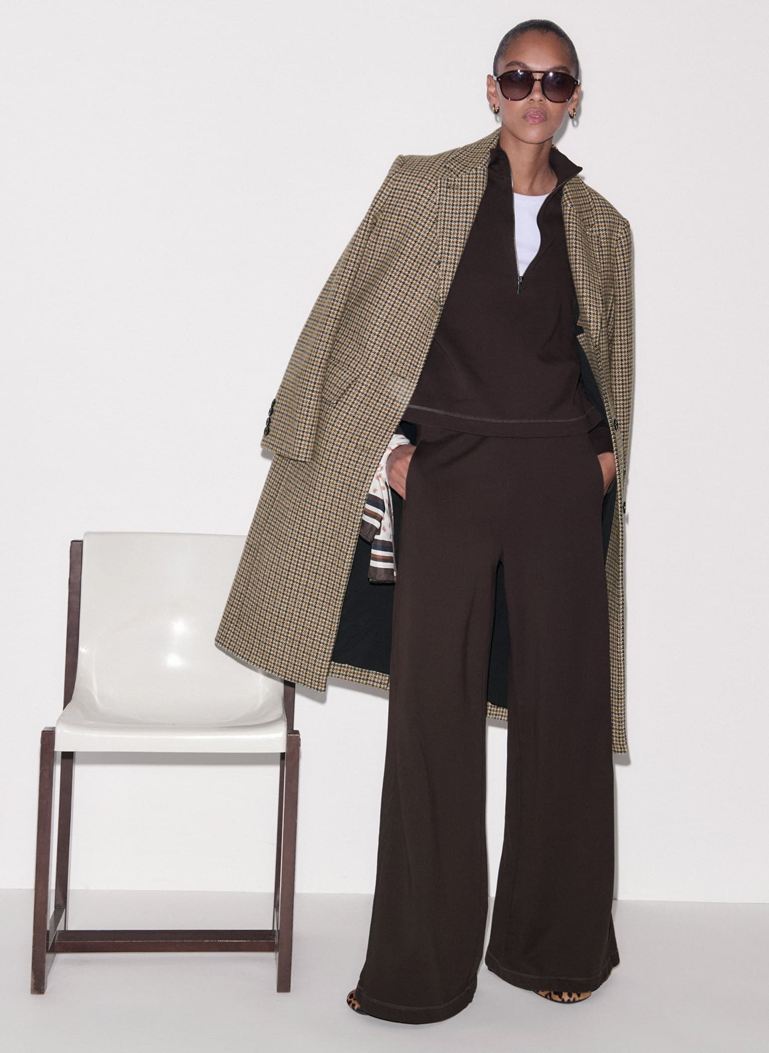 Woman stood up in front of a chair wearing a brown tracksuit and sunglasses, with a checked coat over her shoulders.