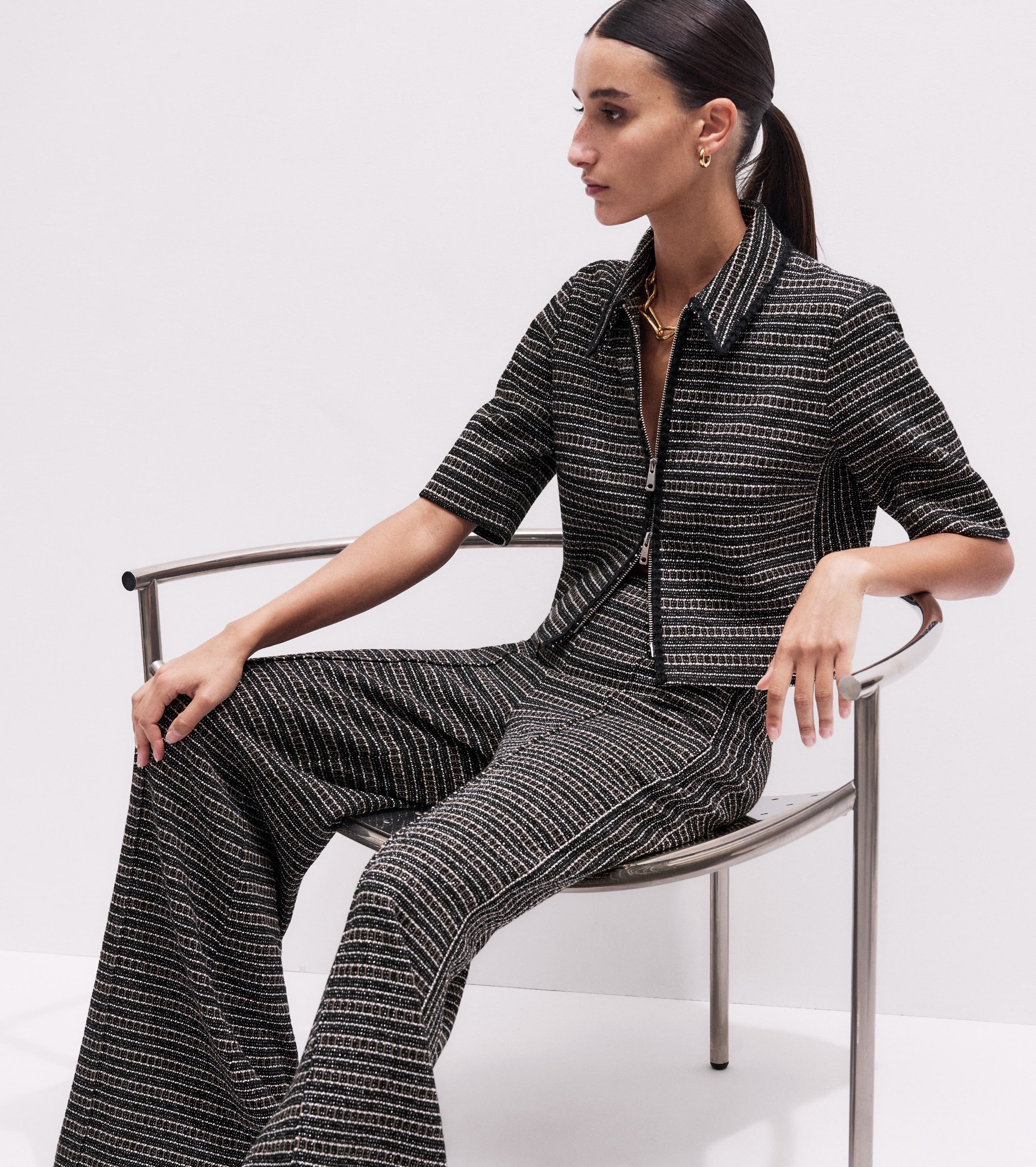 A woman sat in a chair wearing a matching monochrome co-ord outfit and gold jewellery.