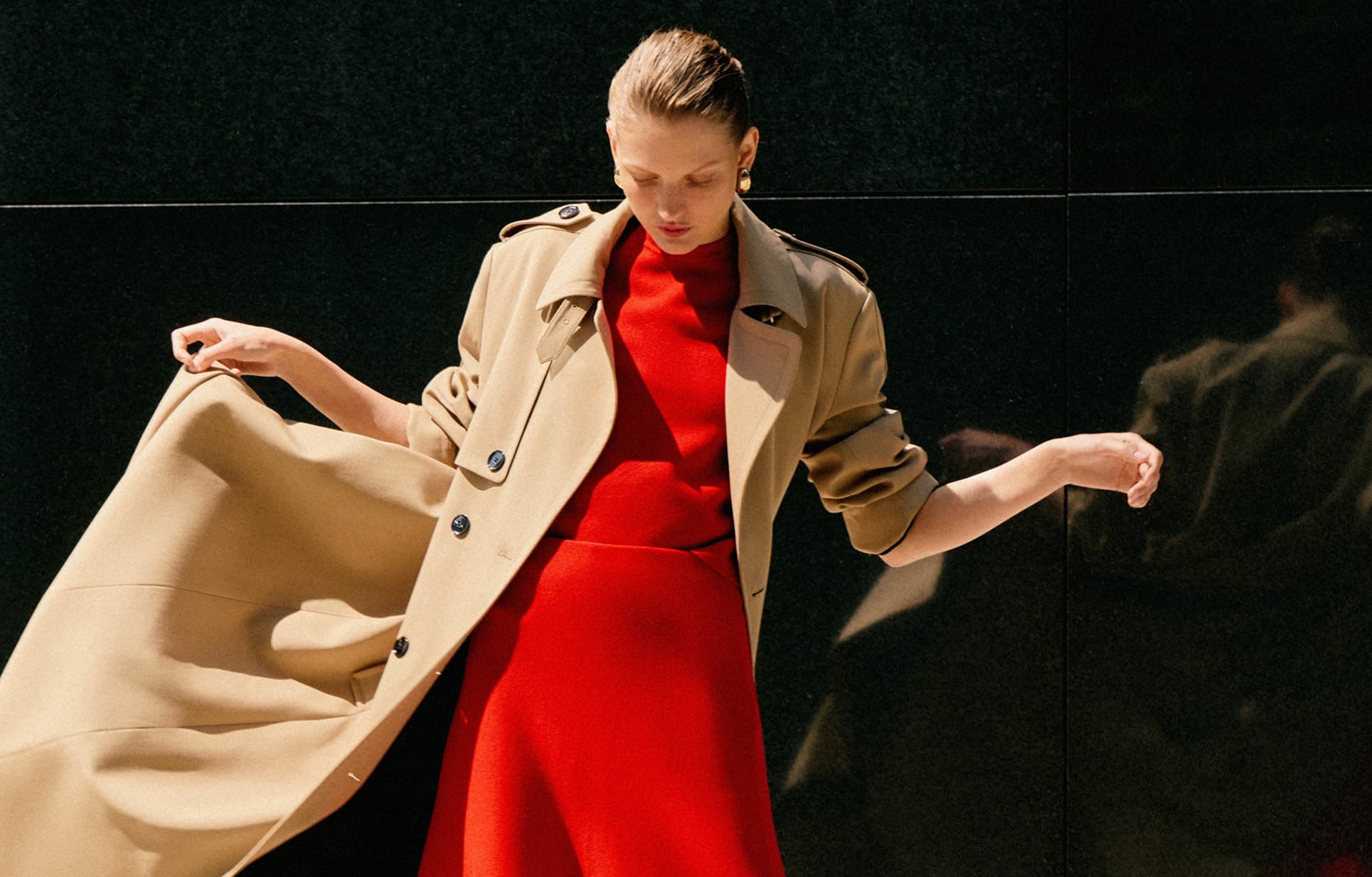 Model wearing matching red skirt and top with a tan trench coat over the top