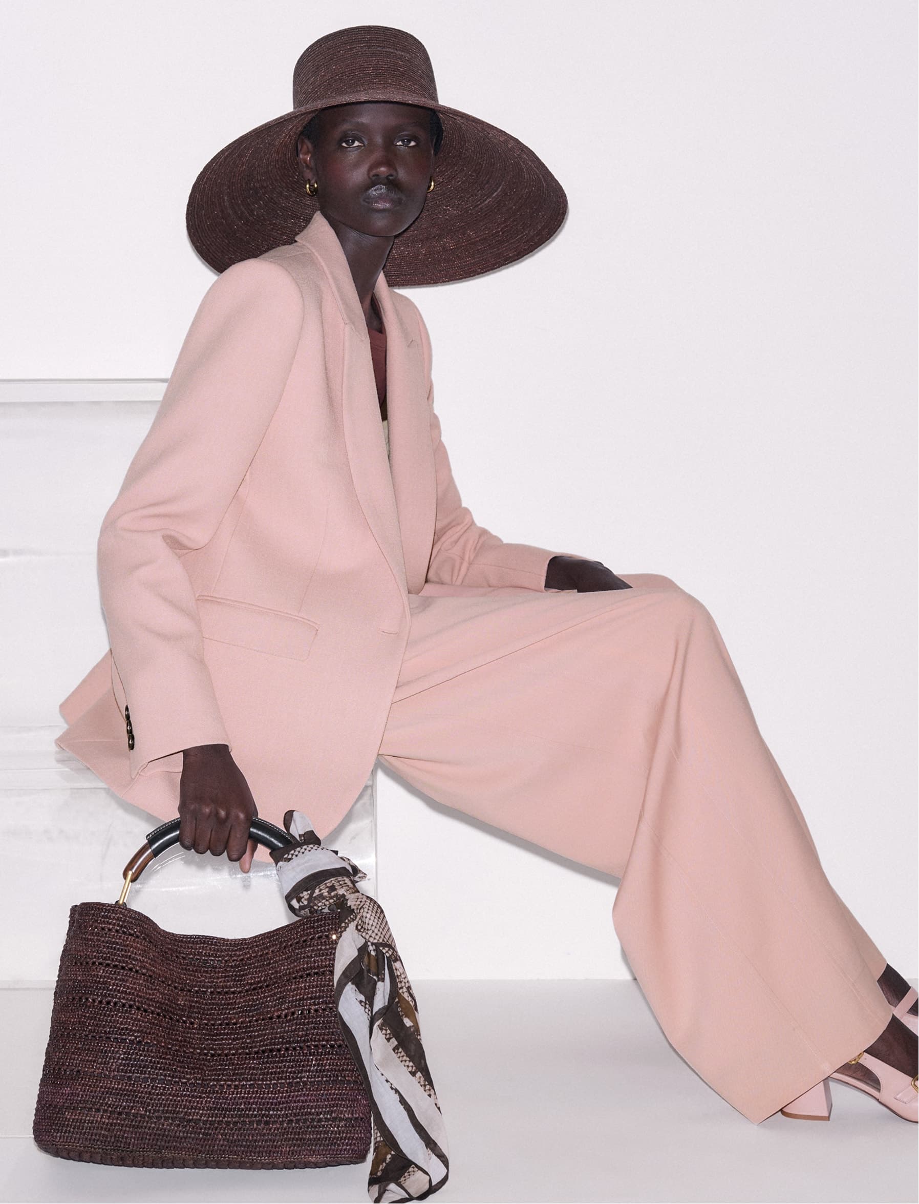 Model sat down in a pink suit, wearing a brown hat and holding a brown raffia bag.