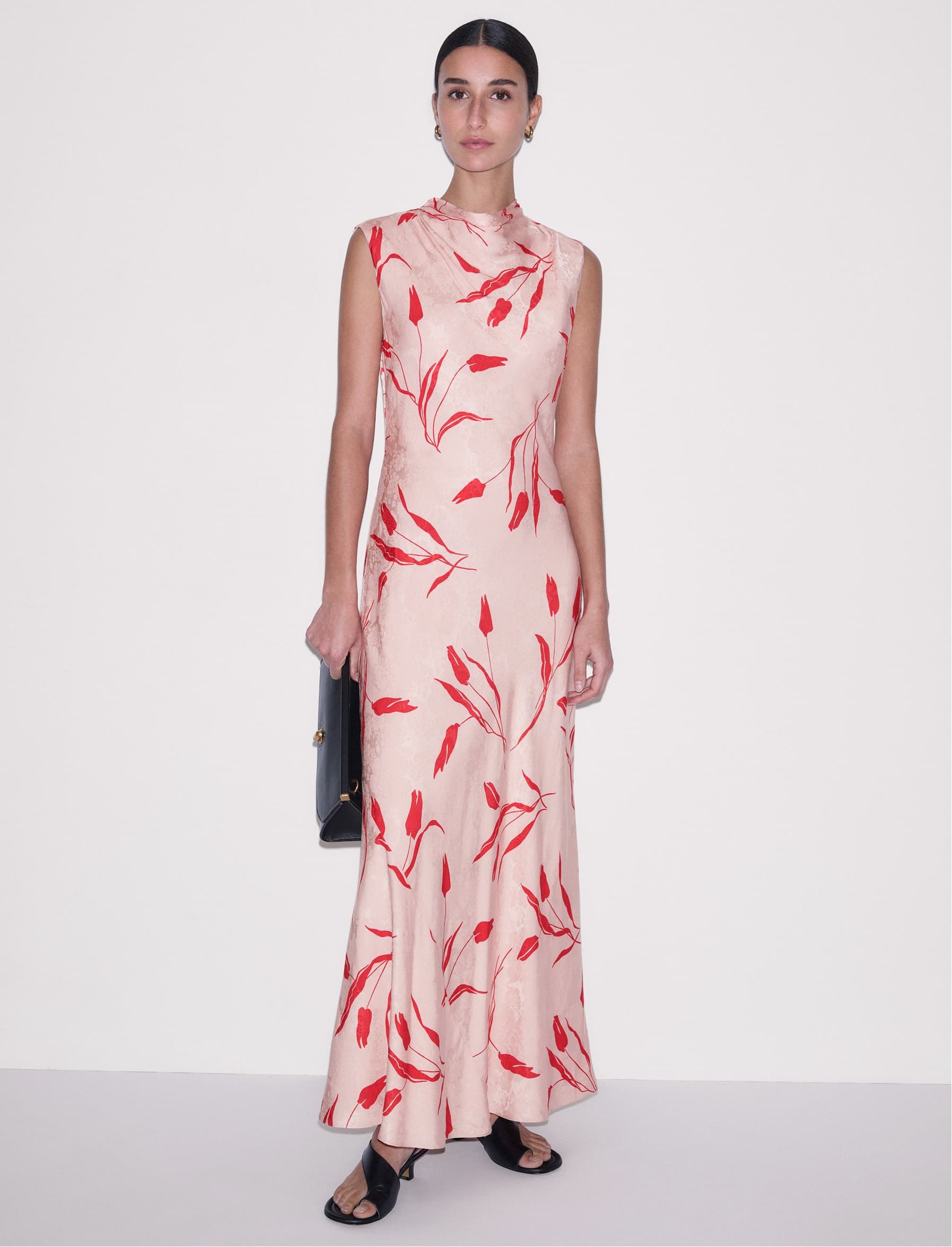 Model standing wearing a pink maxi dress, holding a black clutch bag.