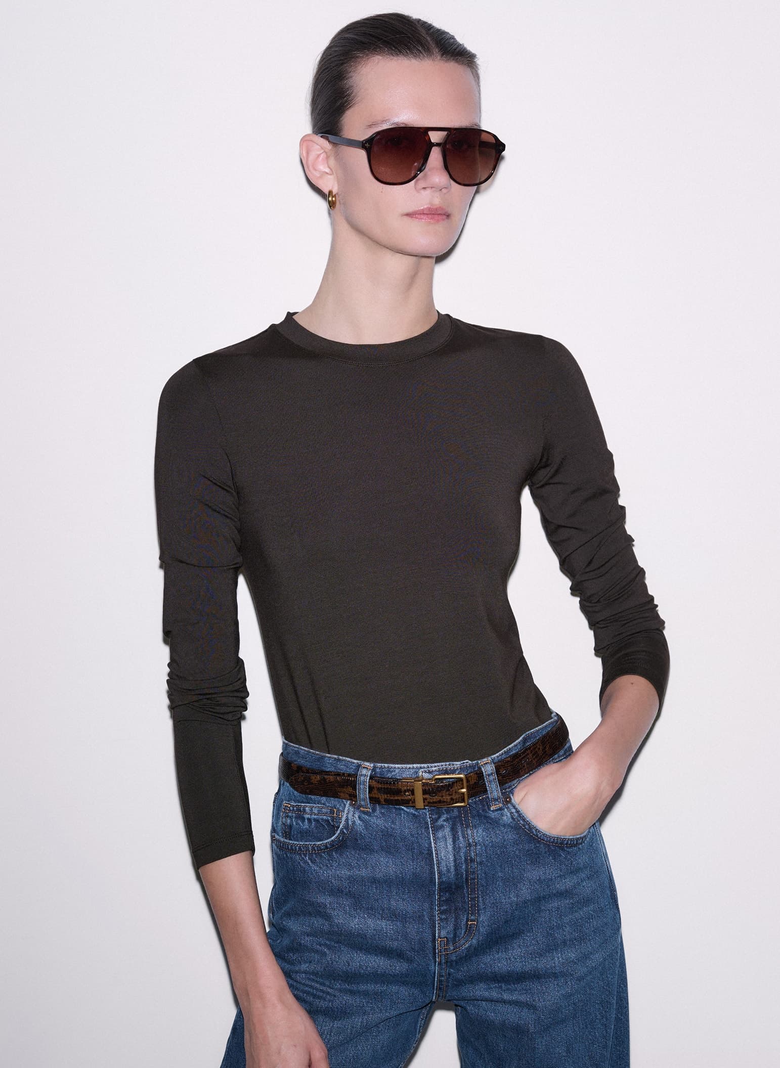 Woman wearing sunglasses and a long-sleeve t-shirt and jeans in front of a plain background.