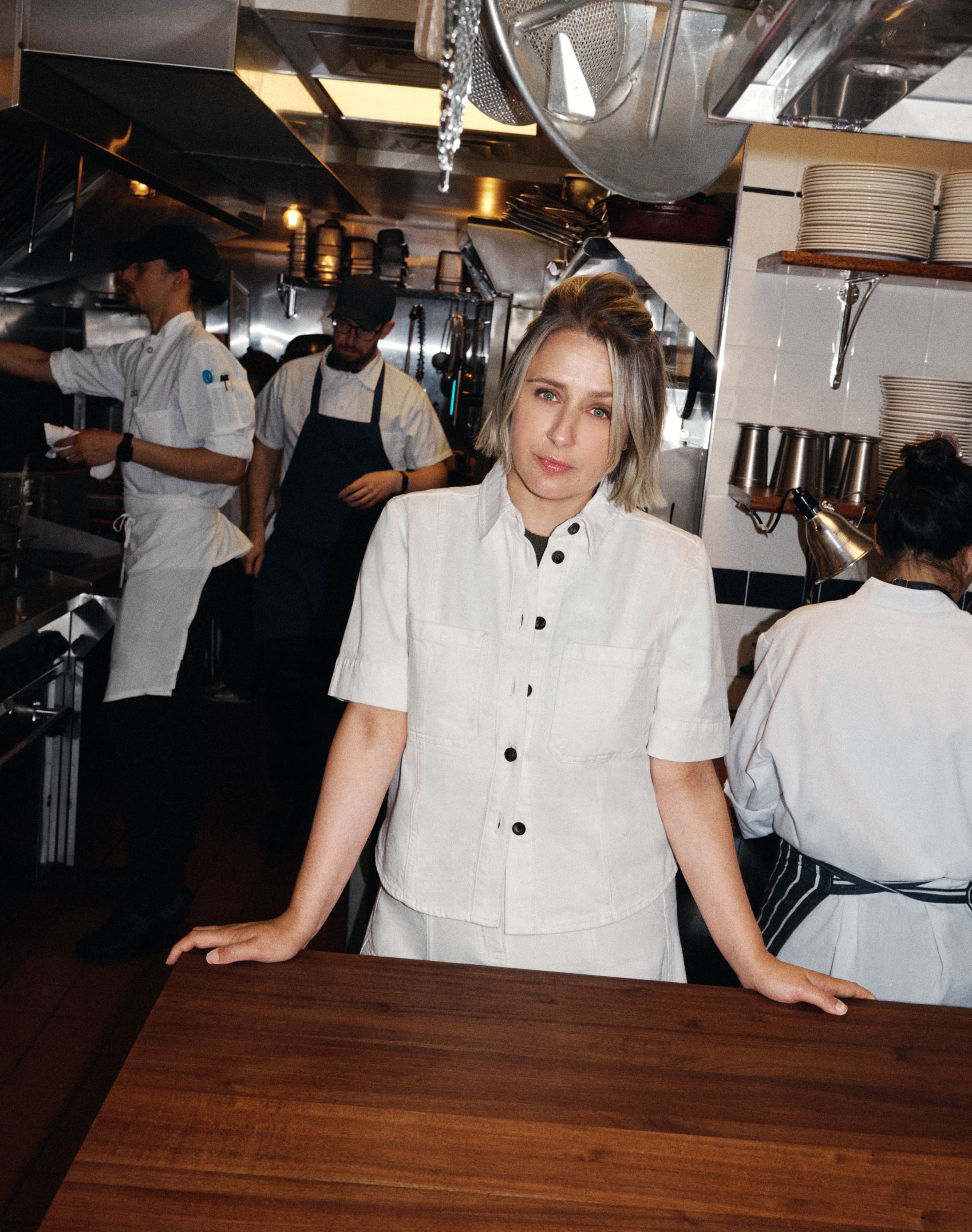 Woman stood in a kitchen with chefs behind her, she is wearing a white button down jacket.