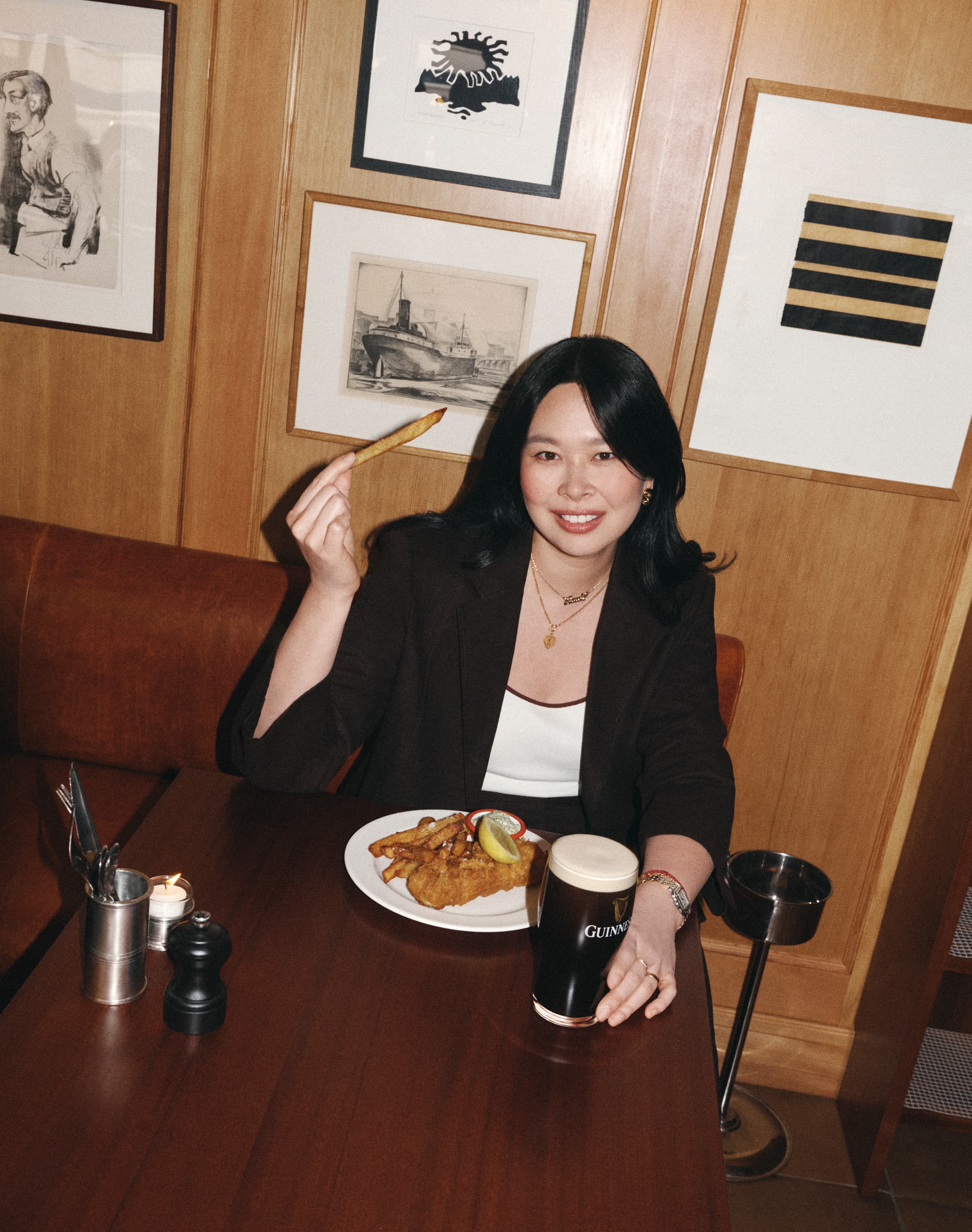 Woman sat at a restaurant table, holding up a chip and enjoying a plate of fish and chips and a pint of guiness.